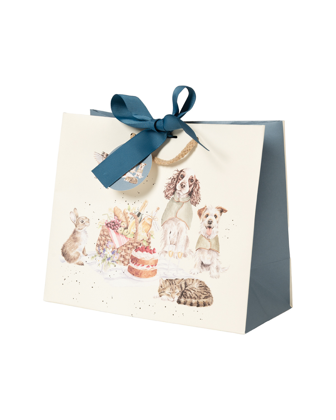 Schrägansicht auf die Geschenktasche "The Hoppy Picnic" von Wrendale Designs. Zu sehen ist eine Illustration von zwei Hunden mit Überwurfdeckchen und ein Kaninchen sitzend auf einer Picknickdecke. Vor der Picknickdecke liegt eine braun-schwarz getigerte schlafende Katze. Ein niedliches Rotkehlchen fliegt über die Picknickdecke hinweg, auf welcher sich ein prall gefüllter Picknickkorb und ein Erbeerkuchen befinden. Das mitgelieferte Band verschließt die Tasche als eine kunstvolle blaue Schleife.