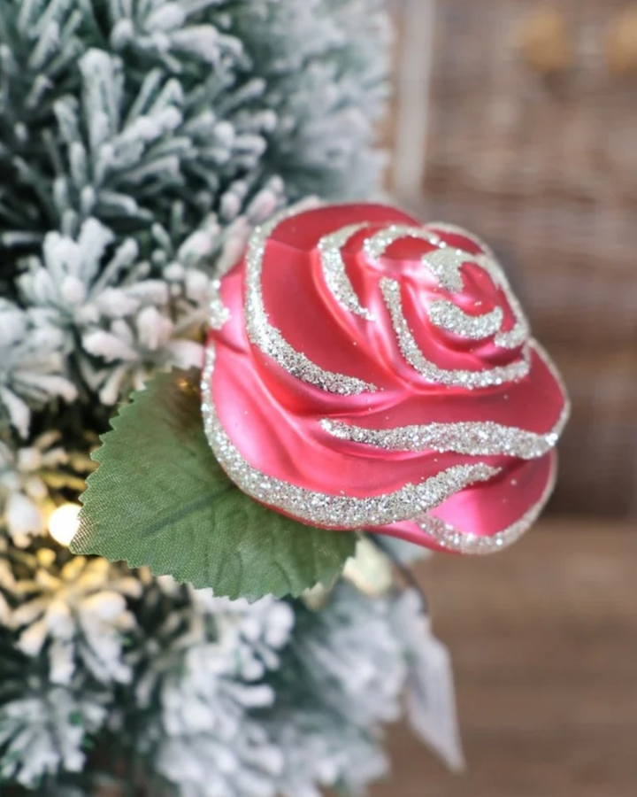 Schräge Ansicht des weihnachtlichen Rosen Ornaments zum Klemmen, welches von Inge Glas in Deutschland handgefertigt wurde. Die Rose hat rote Blütenblätter mit silbernen Glitzer Highlights und einem grünen Blatt unterhalb der Blüte. Auf diesem Bild in einen weiß gepuderten Baum geklemmt.