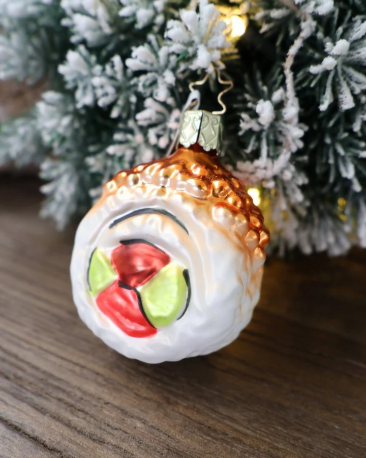 Schräge Ansicht des weihnachtlichen Sushi Ornaments Ura Maki von Inge Glas, welches vor weiß gepudertem Tannengrün auf einem Holztisch dekoriert ist.