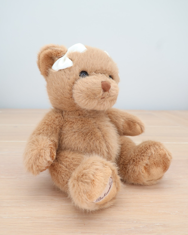 Ein brauner Teddybär von Bukowski mit weißer Schleife im Haar sitzt seitlich auf einem hellen Holztisch.