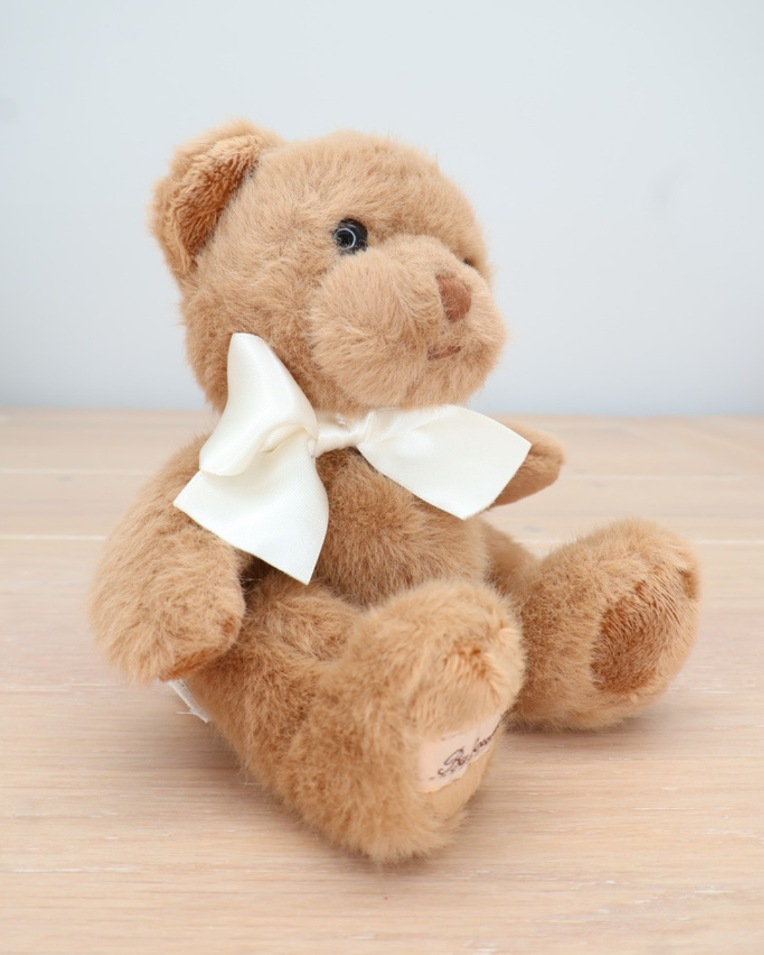Ein brauner Teddybär von Bukowski mit cremefarbener Fliege sitzt auf einem hellen Holztisch und guckt zur Seite.