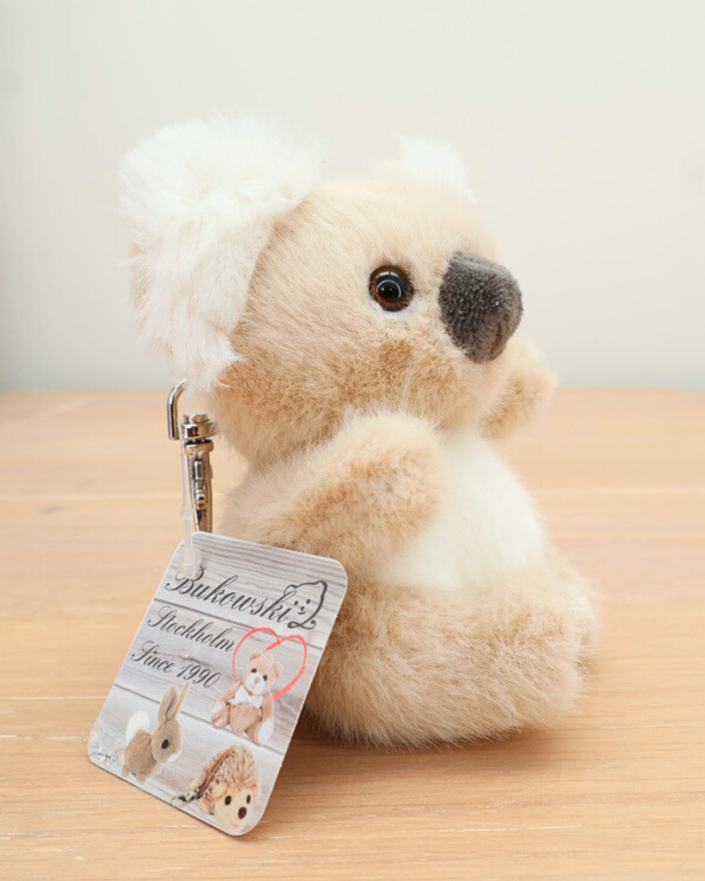 Der Baby-Koala Schlüsselanhänger von Bukowski sitzt auf einem Holztisch. Der Metallkarabiner und das Markenetikett „Bukowski Stockholm Since 1990“ sind gut erkennbar.