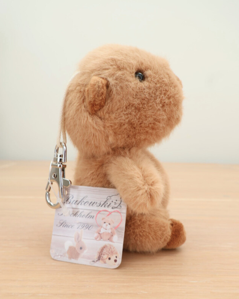 Der Capybara Schlüsselanhänger Theo von Bukowski sitzt auf einem Holztisch. Der Metallkarabiner und das Markenetikett „Bukowski Stockholm Since 1990“ sind sichtbar.