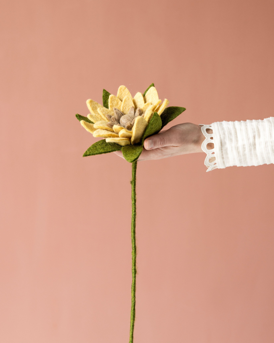 Hand hält eine gelbe Filz-Sonnenblume von Gry & Sif vor rosafarbenem Hintergrund