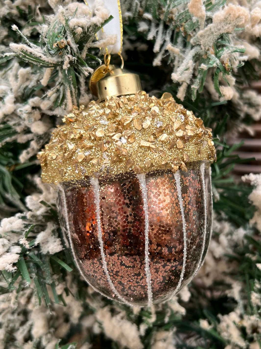 Eichel Weihnachtsbaumanhänger Sparkling Acorn Ornament von Rivièra Maison in braun und gold mit viel Glitzer an einem mit Kunstschnee bedecktem Tannenbaum.