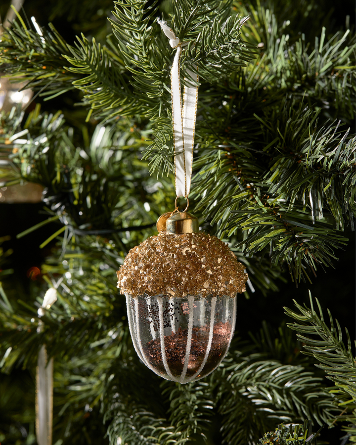 Weihnachtsbaumschmuck „Sparkling Acorn Ornament“ von Rivièra Maison am Tannenzweig. Die gläserne Kugel in Form einer goldglitzernden Eichel reflektiert festlich das Licht.