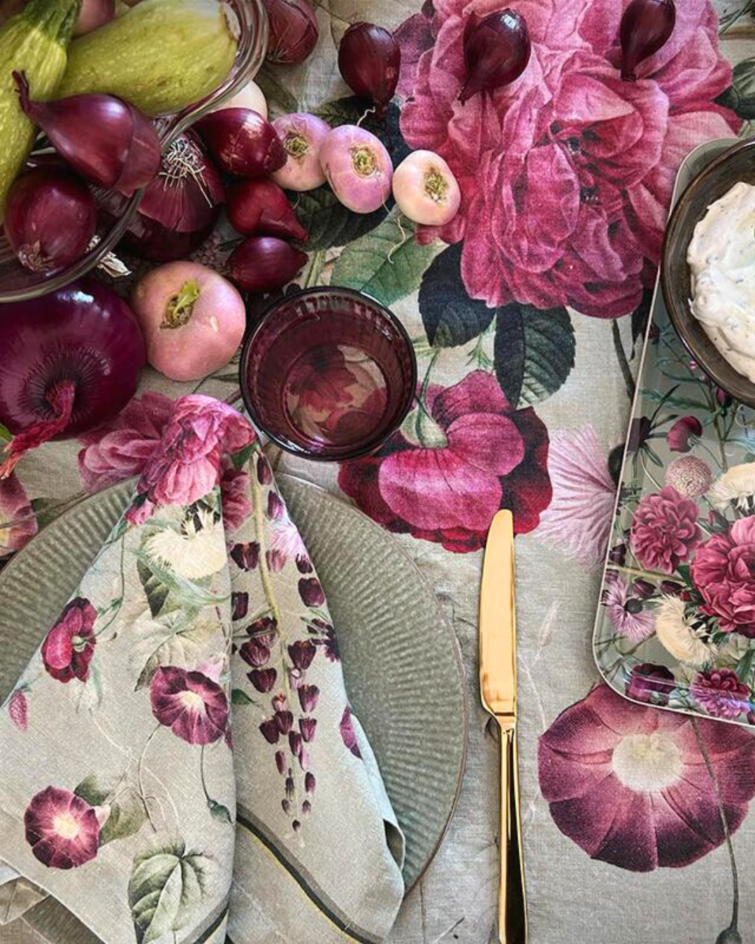 Eine Leinenserviette Blossom Garden von Koustrup & Co., locker auf einem Teller mit goldener Gabel platziert, umgeben von einer Tischplatte mit Blumenmotiv.