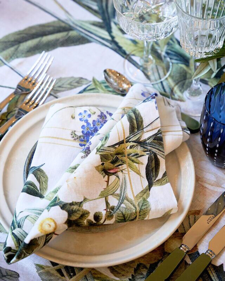 Eine Leinenserviette „Blue Flower Garden“ von Koustrup & Co., locker gefaltet, liegt auf einem weißen Teller, umgeben von Besteck und dekorativen Elementen auf einer festlich gedeckten Tafel.