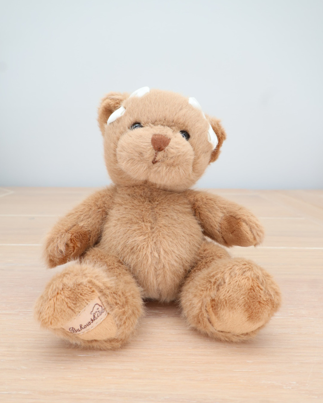 Ein brauner Teddybär von Bukowski mit weißer Schleife im Haar sitzt auf einem hellen Holztisch.