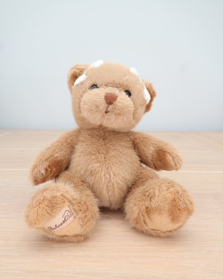 Ein brauner Teddybär von Bukowski mit weißer Schleife im Haar sitzt auf einem hellen Holztisch.