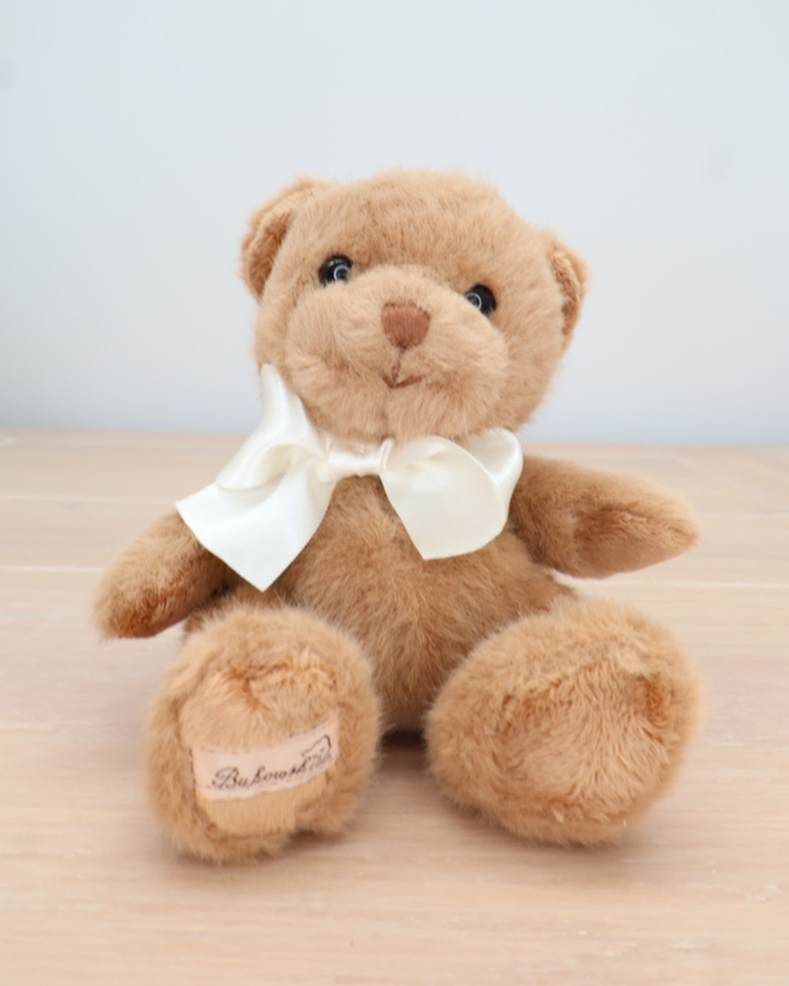 Ein brauner Teddybär von Bukowski mit cremefarbener Fliege sitzt auf einem hellen Holztisch