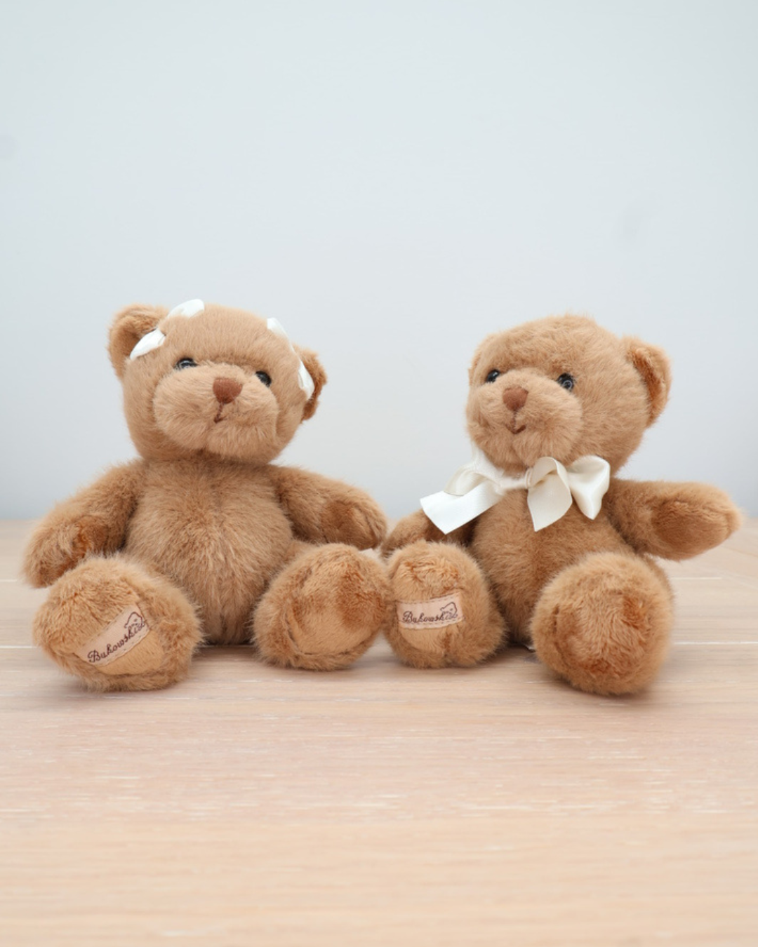Zwei braune Teddybären von Bukowski sitzen auf einem hellen Holztisch. Ein Bär trägt eine Fliege, der andere eine Schleife im Haar.