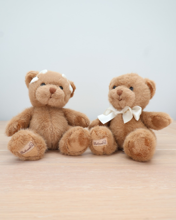 Zwei braune Teddybären von Bukowski sitzen auf einem hellen Holztisch. Ein Bär trägt eine Fliege, der andere eine Schleife im Haar.