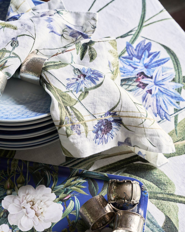 Eine gefaltete Leinenserviette „Blue Flower Garden“ von Koustrup & Co., mit einem dekorativen Metallring gebunden, liegt auf einem Stapel weißer Teller mit floralem Geschirr im Hintergrund.