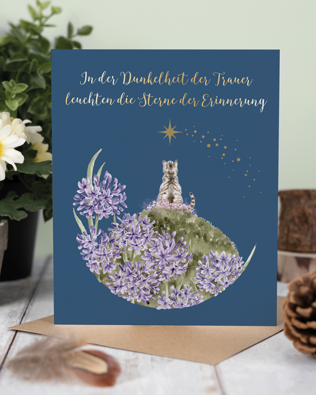 Die Trauerkarte "A Forever Friend" von Wrendale Designs zeigt eine liebevoll von Hannah Dale illustrierte Katze, welche in lila-rosafarbenen Blumen sitzend in Richtung Sternenhimmel aufblickt. Im oberen Drittel der Karte ist in goldenen Lettern der Spruch "In der Dunkelheit der Trauer leuchten die Sterne der Erinnerung" aufgebracht. Hier auf einem Tisch stehend mit Feder, Tannenzapfen und Blumenvase als Dekoration auf einek weißen Holztisch.