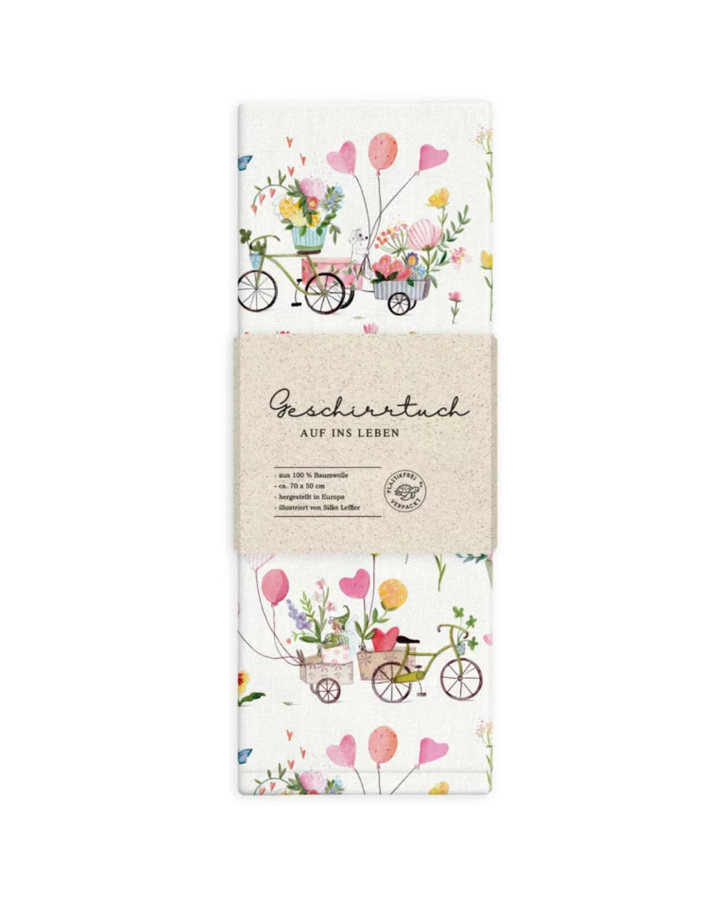 Gefaltetes Geschirrtuch „Auf ins Glück“ vom Grätz Verlag mit Papierbanderole und sichtbarem Fahrrad- und Blumenmotiv, Design von Silke Leffler.
