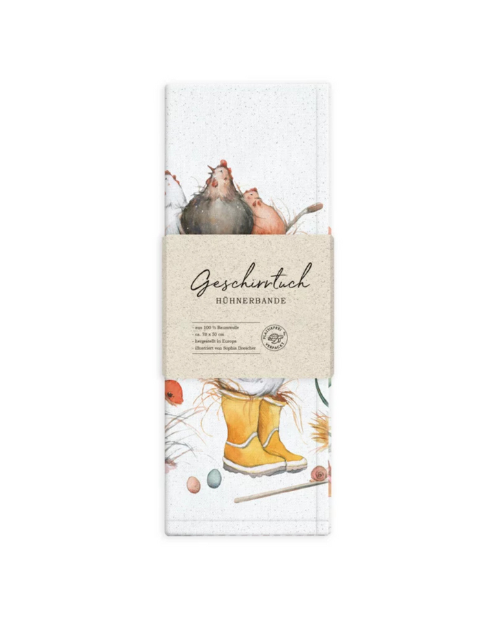 Nahansicht der Verpackung vor weißem Hintergrund mit sichtbarem Teil des Motivs – Hühner in einer Schubkarre, gelbe Gummistiefel, Küken und bunte Eier. Die braune Papierbanderole trägt den Schriftzug „Geschirrtuch Hühnerbande“ und die Angaben zu Größe und Material.