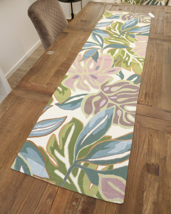 Vollansicht auf den Zimmerpflanzen Tischläufer Selva von Sander Table + Home auf einem braunen Holztisch. Die mehrfarbigen Monsterblätter des Tischläufers sind sich gegenseitig überlappend dargestellt. Blaue, grüne und roséfarbene Blätter mit sich farblich absetzender Blattumrandung sind in den Stoff des Tischläufers gewoben.