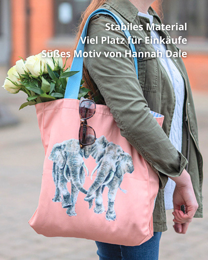 Pinke Elefanten Einkaufstasche von Wrendale Designs. Das Motiv beinhaltet 2 Elefanten welche mit ihren Rüsseln Händchen halten. Die Tasche wird von einer Frau getragen, an ihr hängt eine Sonnenbrille und sie ist gefüllt mit weißen Tulpen.