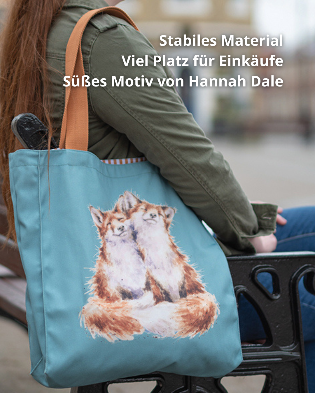 Blaue Fuchs Einkaufstasche von Wrendale Designs getragen von einer Frau welche auf einer Bank sitzt. Zu sehen sind 2 niedlichen kuschelnden Füchse. Der Griff der Tasche ist braun.