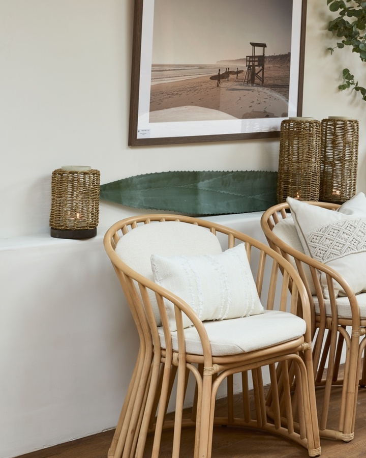 Windlicht "Palmanova Hurricane" von Riviera Maison dekoriert auf einem Sims vor einer weißer Wand. Zwei Holzstühle mit weißen Kissen stehen im Vordergrund. An der Wand hängt ein Foto mit Strandmotiv und Surfern.