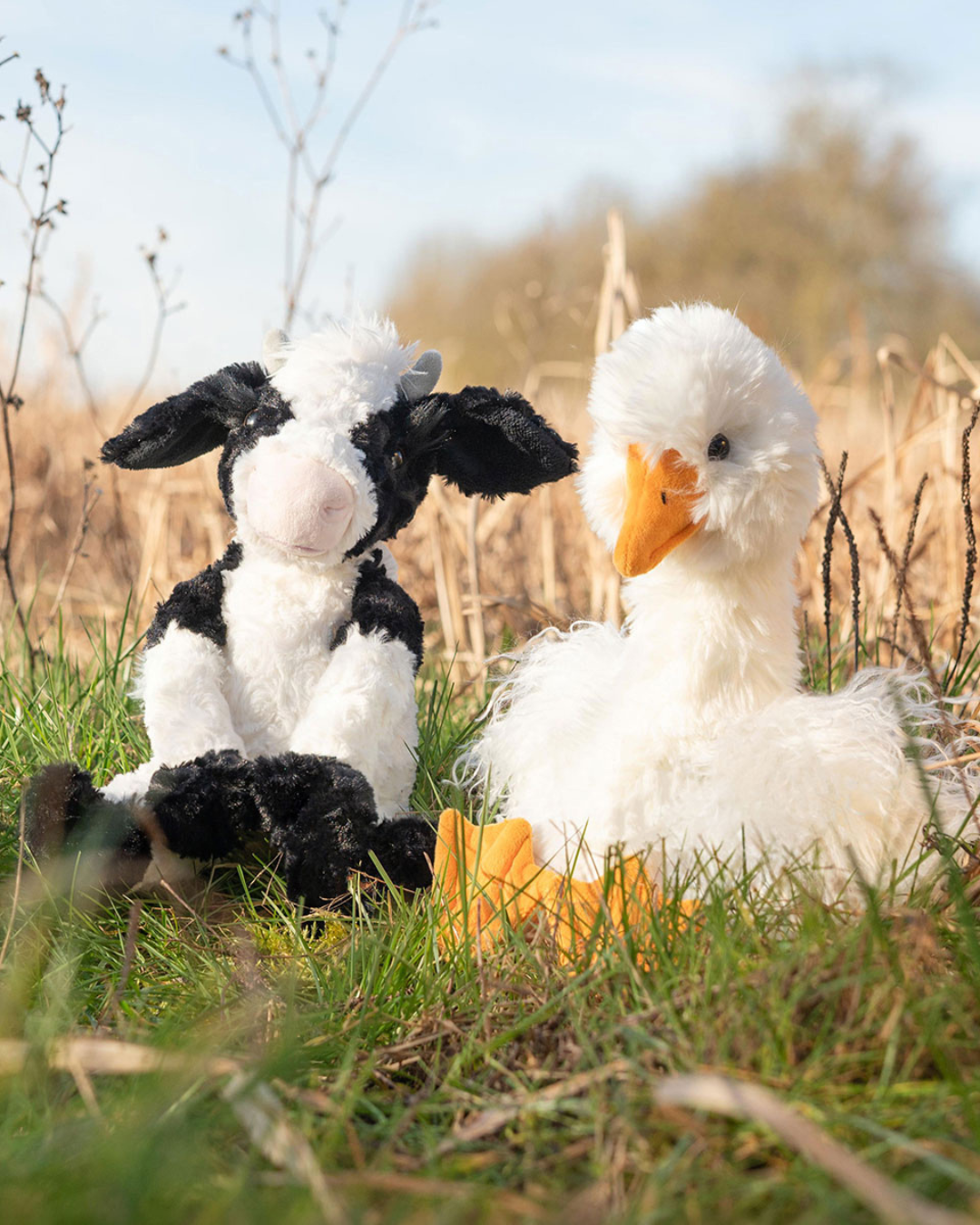 Gans Kuscheltier "Pearl" und Kuh Kuscheltier "Mathilda" sitzen nebeneinander auf einer Wiese. Im Hintergrund sind getrocknete Pflanzen erkennbar.