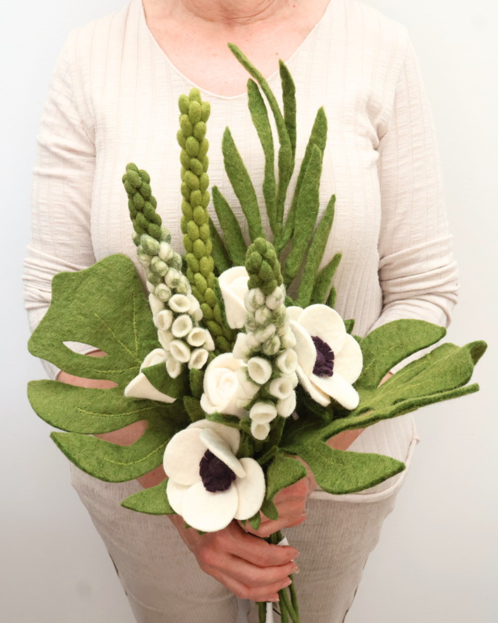 Hochzeits-Blumenstrauß aus Filz in einer Hand gehalten, umgeben von Palmblättern und einem hellen Tonuntersetzer.