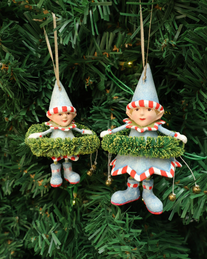 Zwei Größen der "Dashers Elf" Krinkles von Patience Brewster hängen an einem Tannenbaum. Zu sehen sind 2 Elfen mit hellblauen Kleidern, Hüten, Schuhen und grünen Pflanzen Details. Die Elfen halten 2 Glocken in den Händen.
