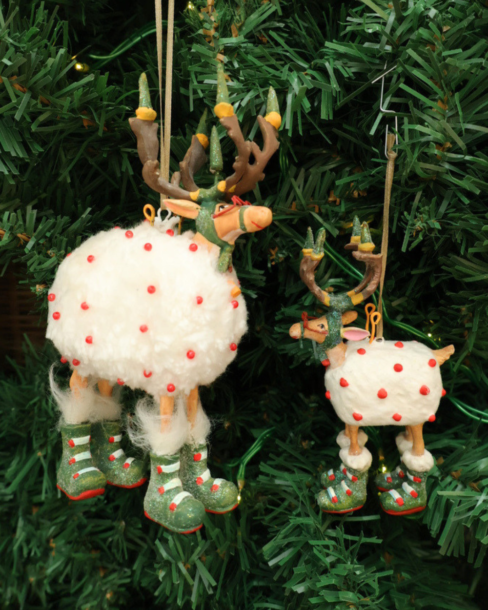 Zwei Größen der "Dash Away Blitzen Rentier" Krinkles von Patience Brewster hängen an einem Tannenbaum. Zu sehen sind 2 Rentiere mit rot bepunkteten Schafsfell. Die Rentiere haben viele grüne und rote Details.