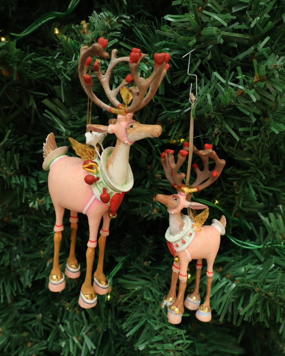 Zwei Größen der "Dash Away Cupid Rentier" Krinkle von Patience Brewster hängen an einem Tannenbaum . Zu sehen sind 2 farbenfrohe Rentier Krinkle mit vielen pinken und roten Details.