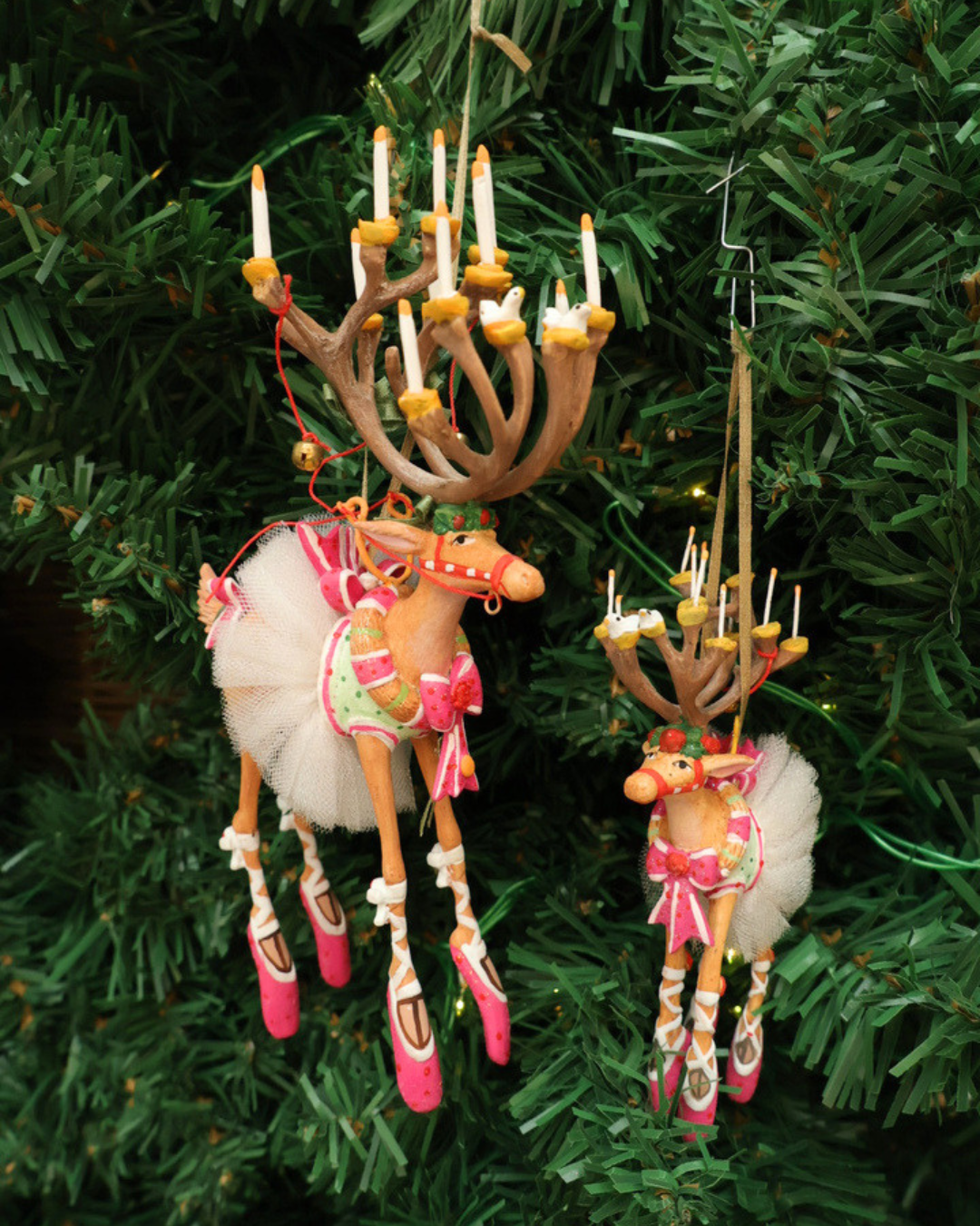 Zwei Größen der "Dash Away Dancer Rentier" Krinkle von Patience Brewster hängen an einem Tannenbaum. Zu sehen sind 2 farbenfrohe Rentier Krinkle welche weiße Röcke tragen mit vielen pinken und weißen Details.