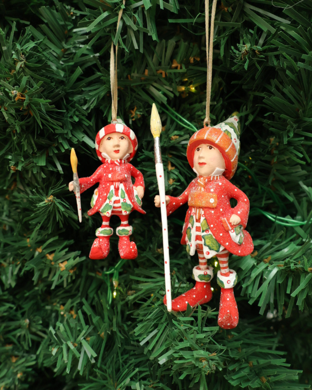 Zwei Größen der "Vixens Elf" Krinkles hängen an einem Tannenbaum. Zu sehen sind 2 größtenteils rot bekleidete Elfen mit weißen Pünktchen und pflanzlichen Details und großen Pinseln in den Händen. 