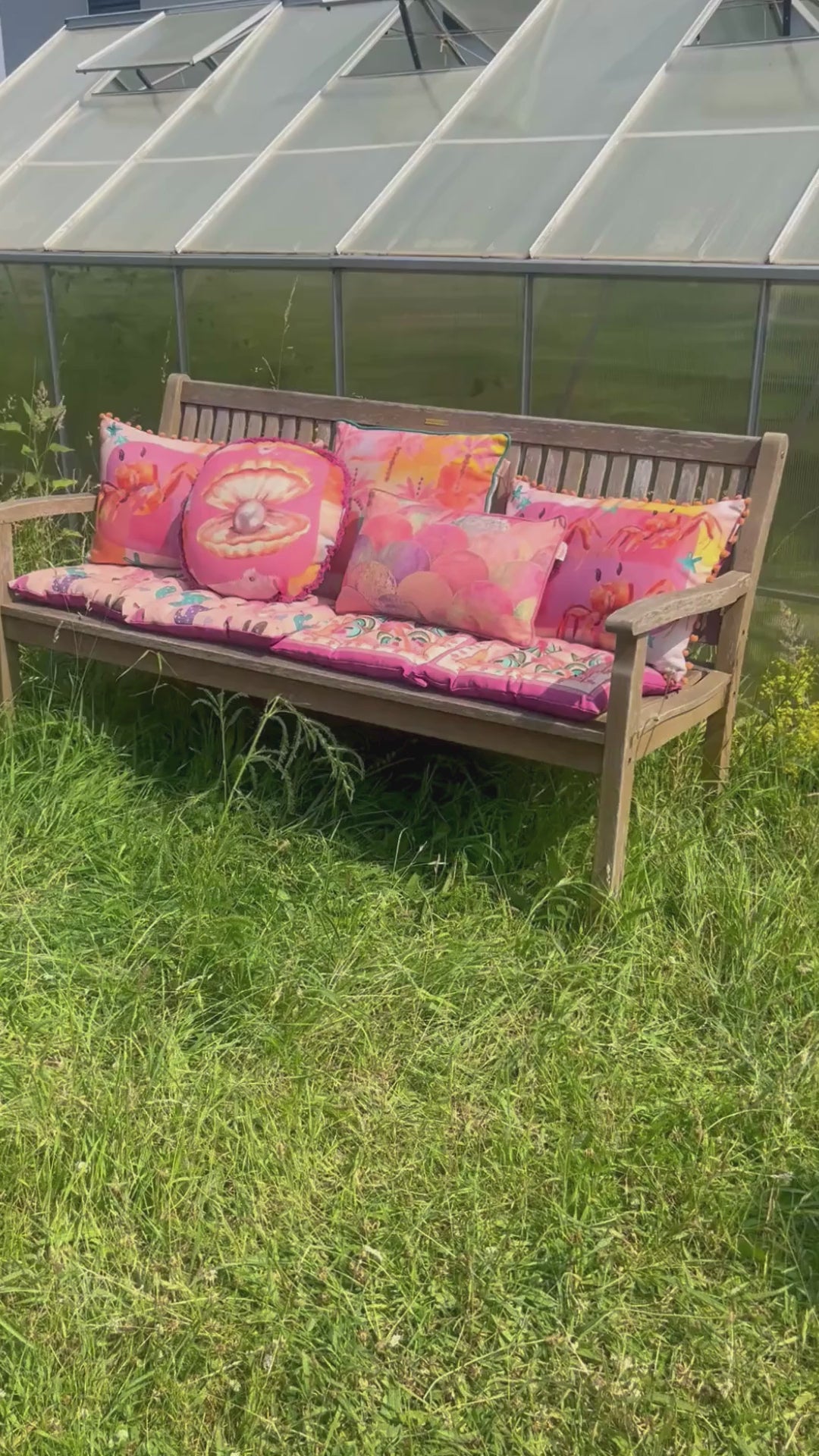 Pinke Outdoor Kissen auf einer Bank im Grünen vor einem Gewächshaus dekoriert.