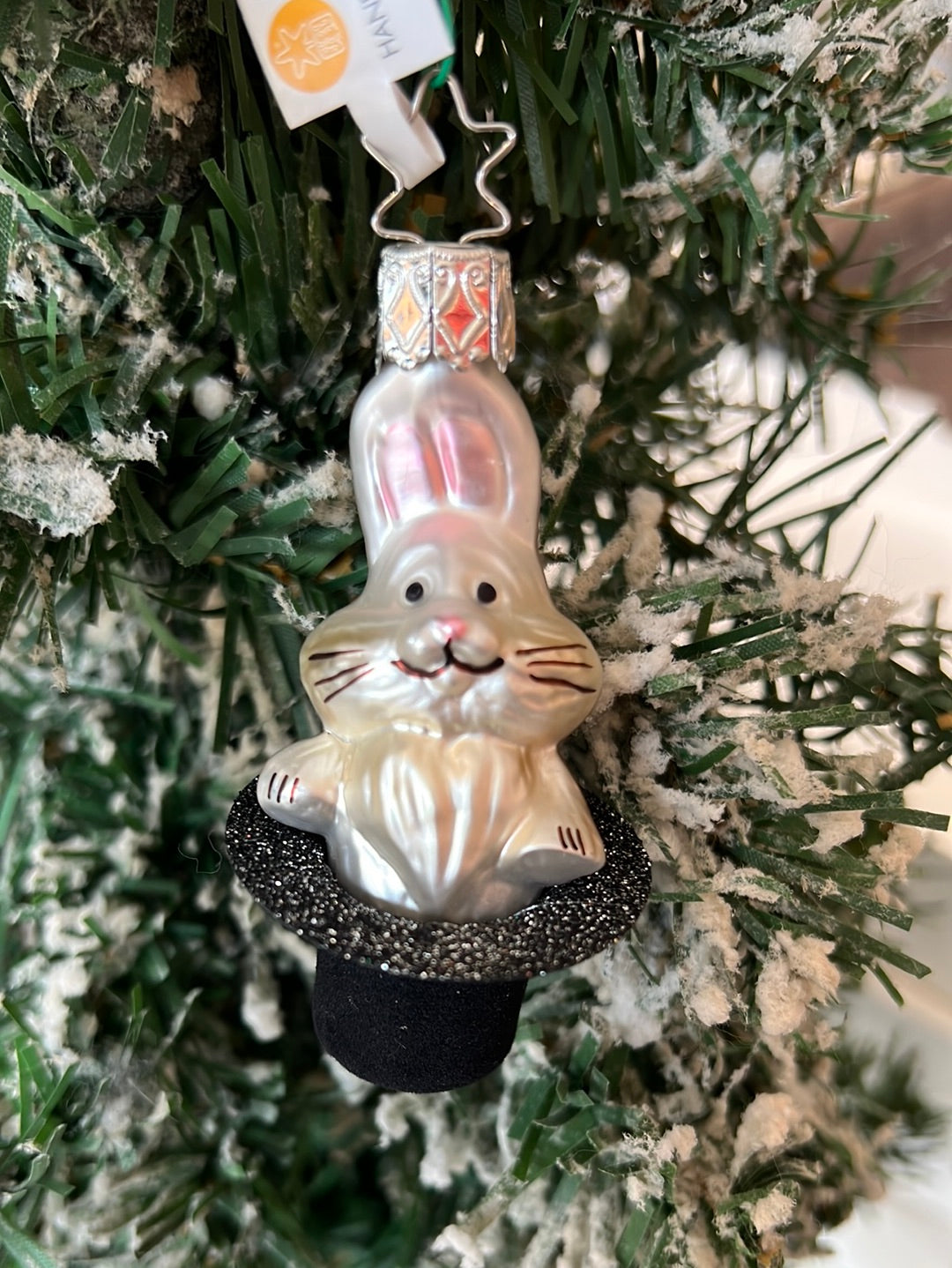 Handgefertigtes Hase im Hut Ornament von Inge Glas. Ein weißer Hase schaut verschmitzt auf einem dunklen Zylinder mit Glitzer an der Unterseite der Hutkrempe.
