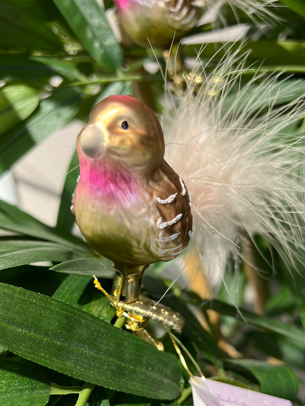 Handgefertigtes Vogelornament "Lotte" zum Klemmen von Inge Glas. Der bräunliche Vogel hat eine hell rosafarbene Brust und silberne Glitzer Highlights an den Flügeln. Echte weiße Federn Formen seinen Schwanz.